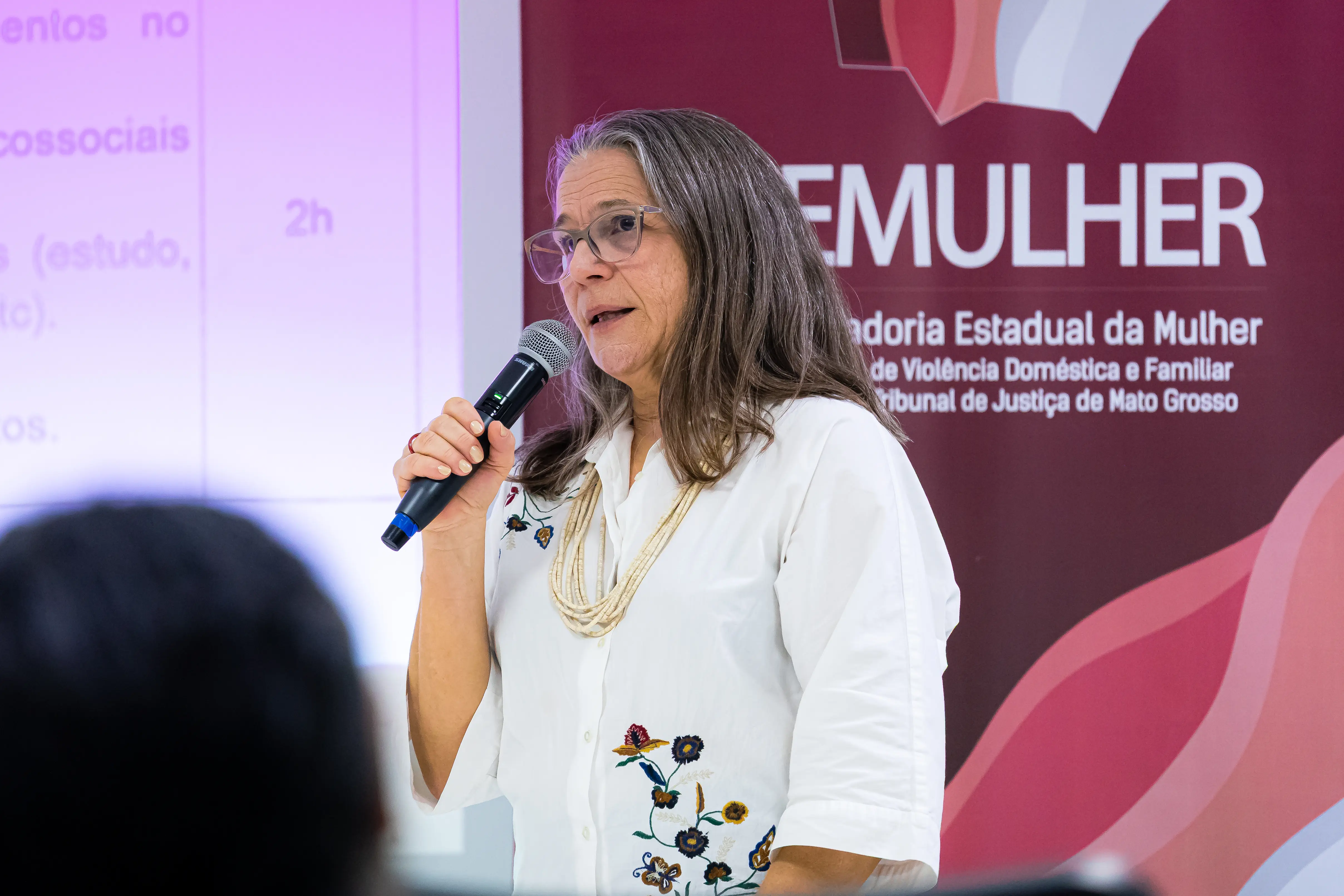 Mulher de óculos e cabelos grisalhos longos fala ao microfone. Ela veste camisa branca com bordados no ombro e colar claro. Ao fundo, um banner vermelho onde se lê 