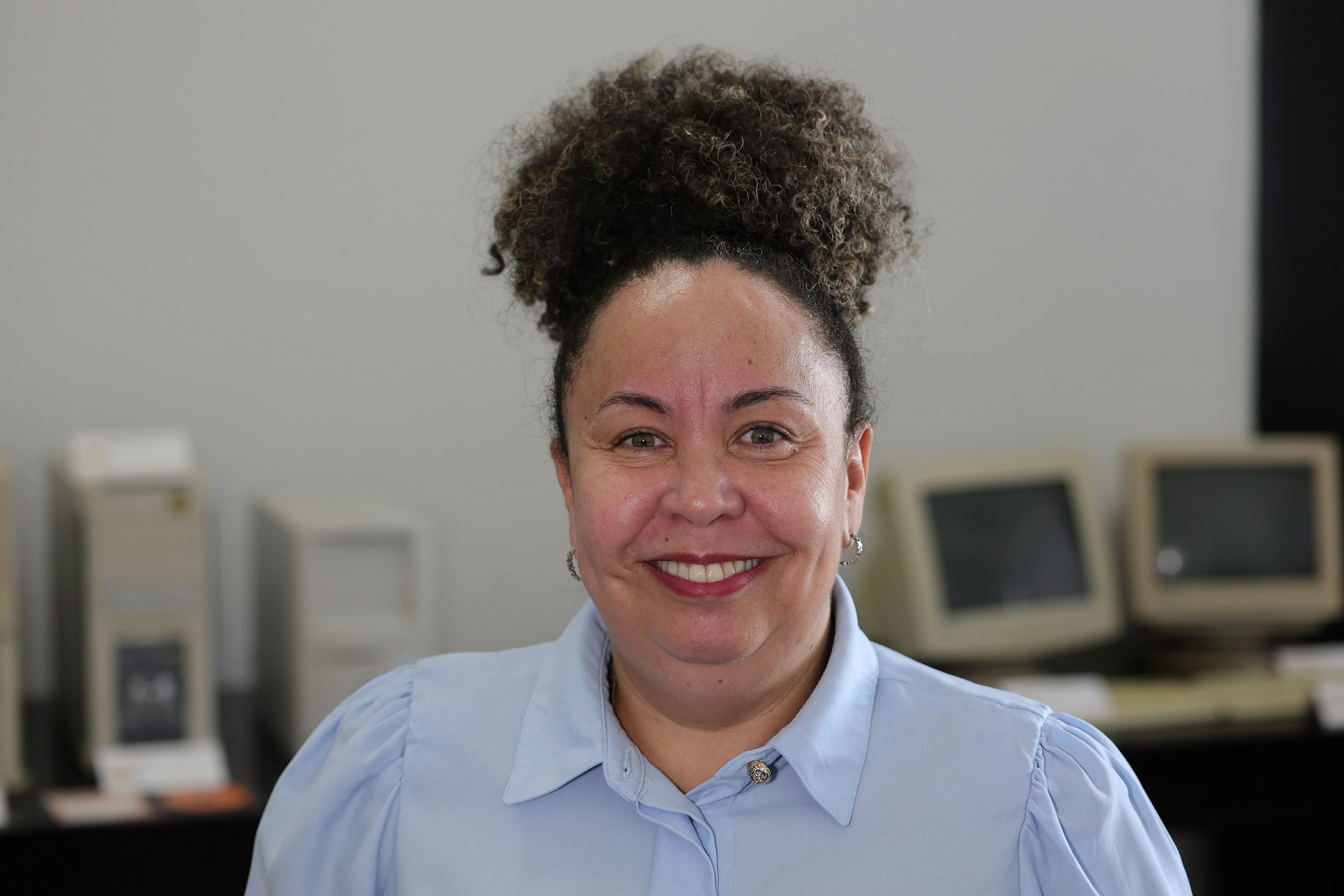 Superintendente do Arquivo Público, Vanda da Silva, é uma mulher de pele clara sorridente, com cabelo crespo escuro preso no topo da cabeça. Ela usa blusa azul-clara de botões. Ao fundo desfocado, há paredes claras e antigos monitores e gabinetes de computador sobre uma bancada escura.