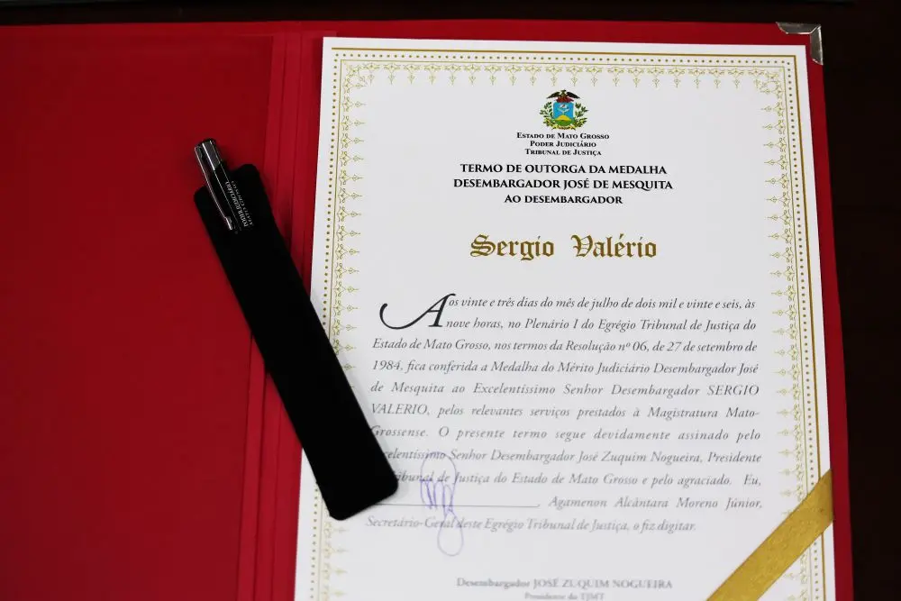 Foto horizontal que mostra um documento e uma cante sobre uma pasta de veludo vermelha. Trata-se do termo que condecorou o desembargador Sérgio Valerio com a medalha do mérito judiciário.