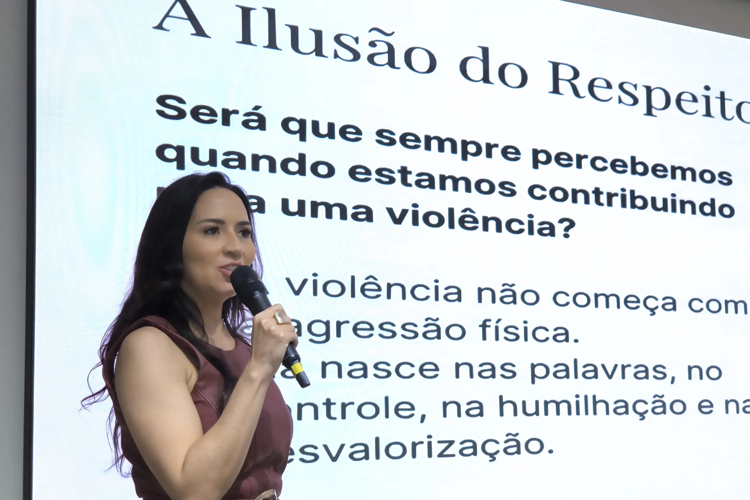 Mulher de cabelos escuros, blusa vinho, fala ao microfone. Ao fundo, telão exibe slide sobre violência doméstica, com o título