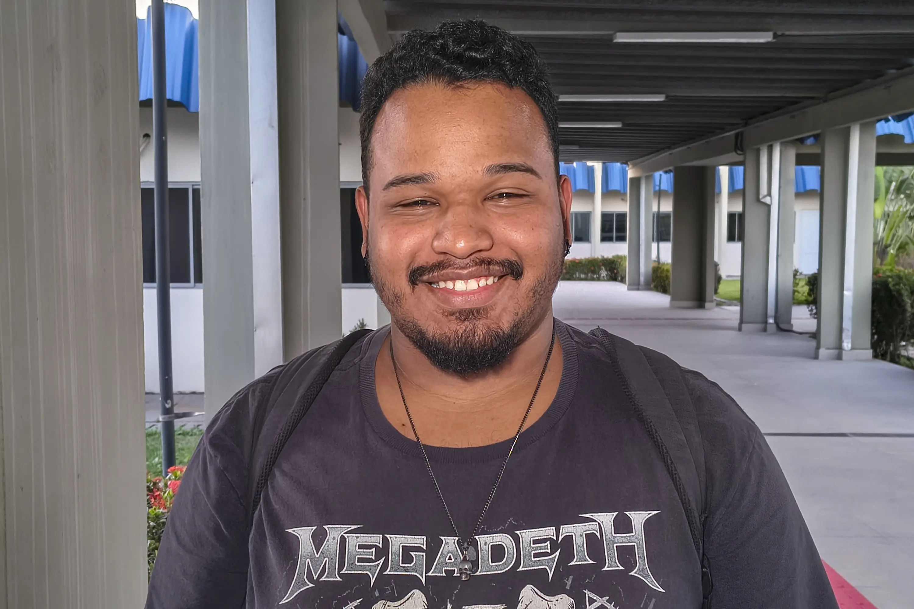 Jovem de barba e cabelos escuros sorri, vestindo camiseta preta estampada e mochila nos ombros. Ao fundo, corredor coberto de prédio com colunas e jardim.
