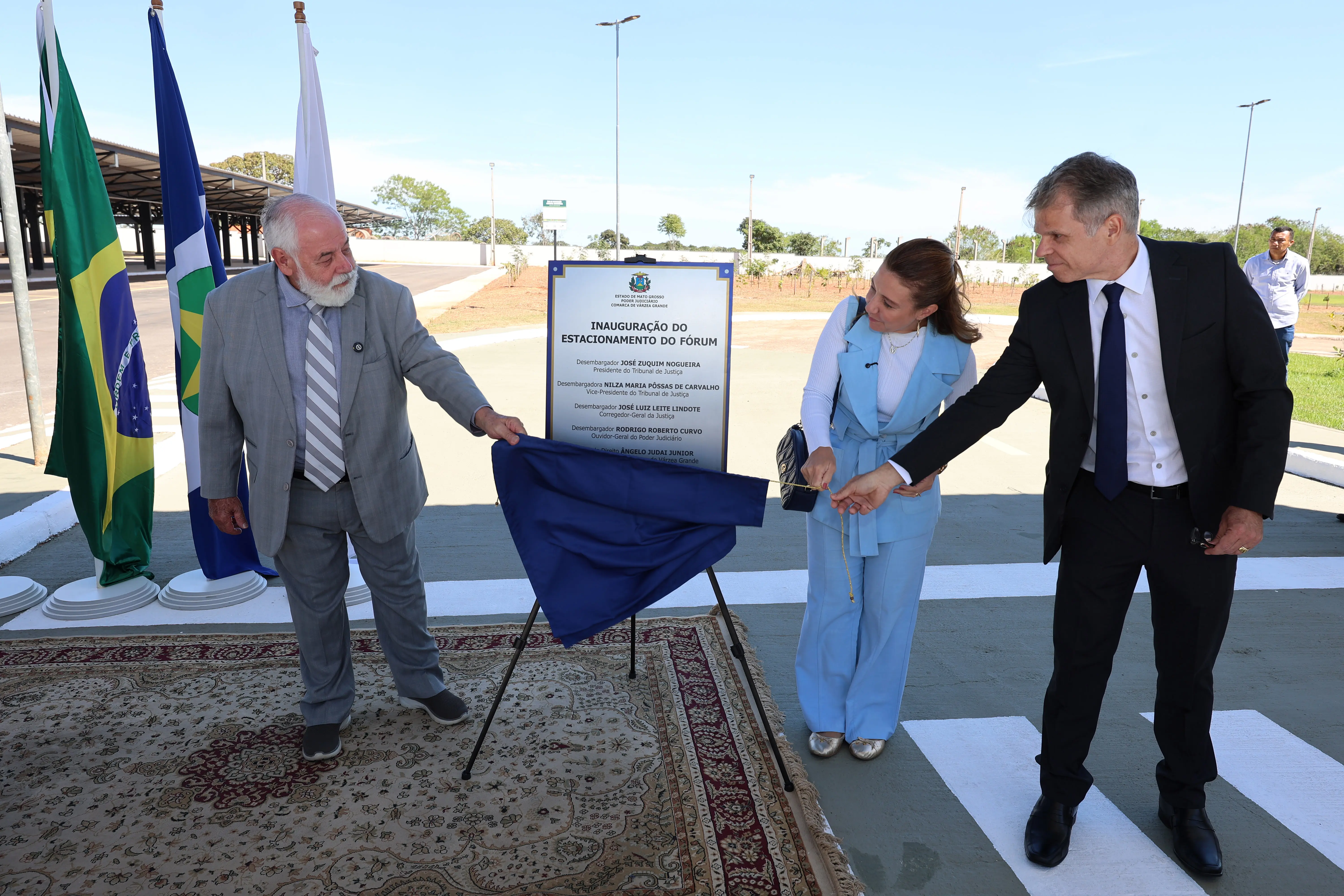 Três pessoas inauguram uma placa descerrando um pano azul em ambiente externo. À esquerda, um homem idoso de terno cinza observa a placa, que traz o texto