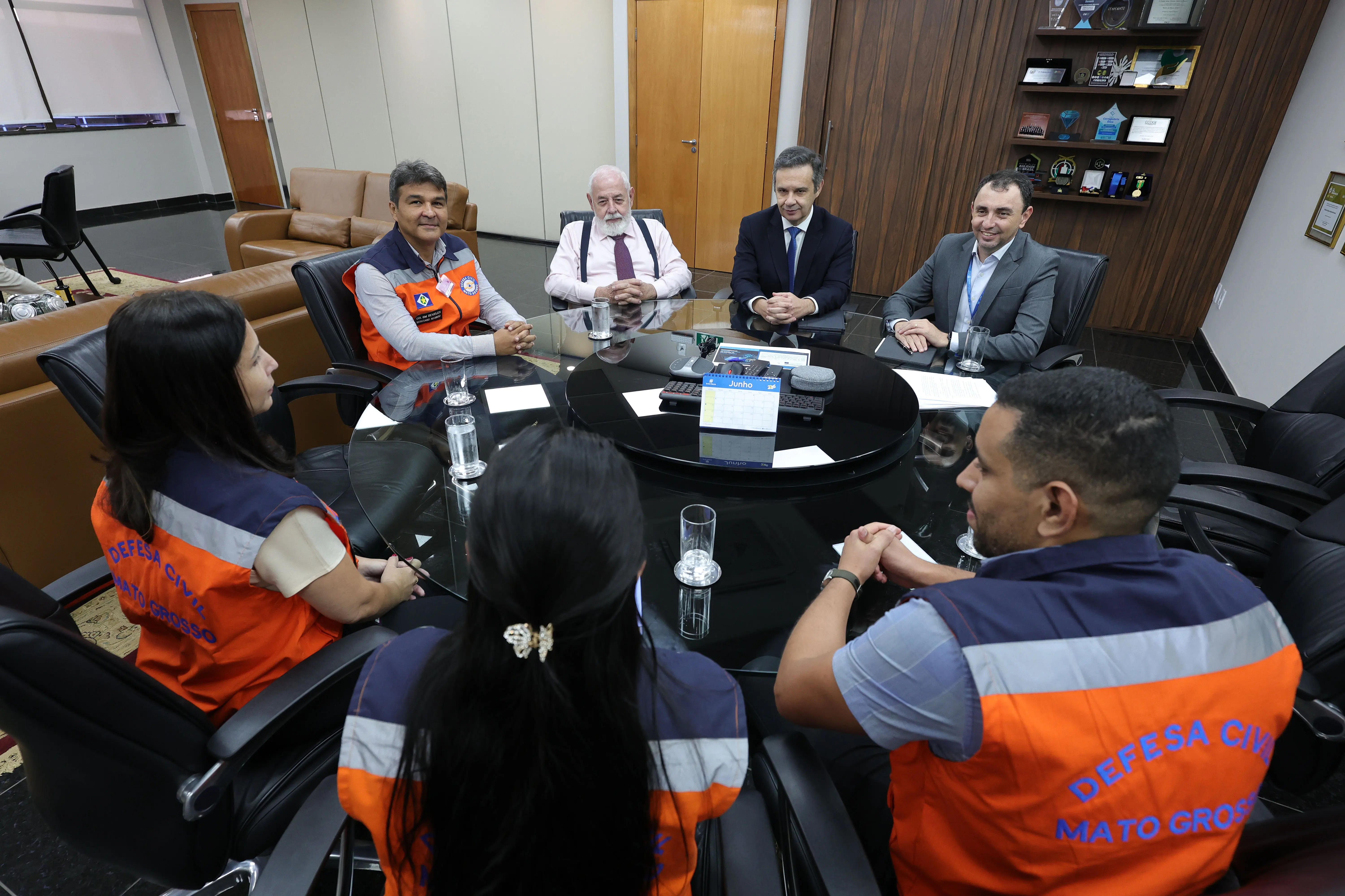 : Sete pessoas reunidas ao redor de uma grande mesa redonda de vidro escuro em um escritório. Algumas usam coletes laranja da Defesa Civil de Mato Grosso e outras vestem trajes formais