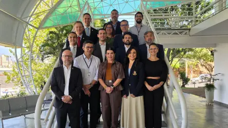 Grupo de pessoas posa em estrutura metálica com cobertura translúcida. Vestem roupas formais, em pé e alinhadas. Ao fundo, vegetação e luz natural.