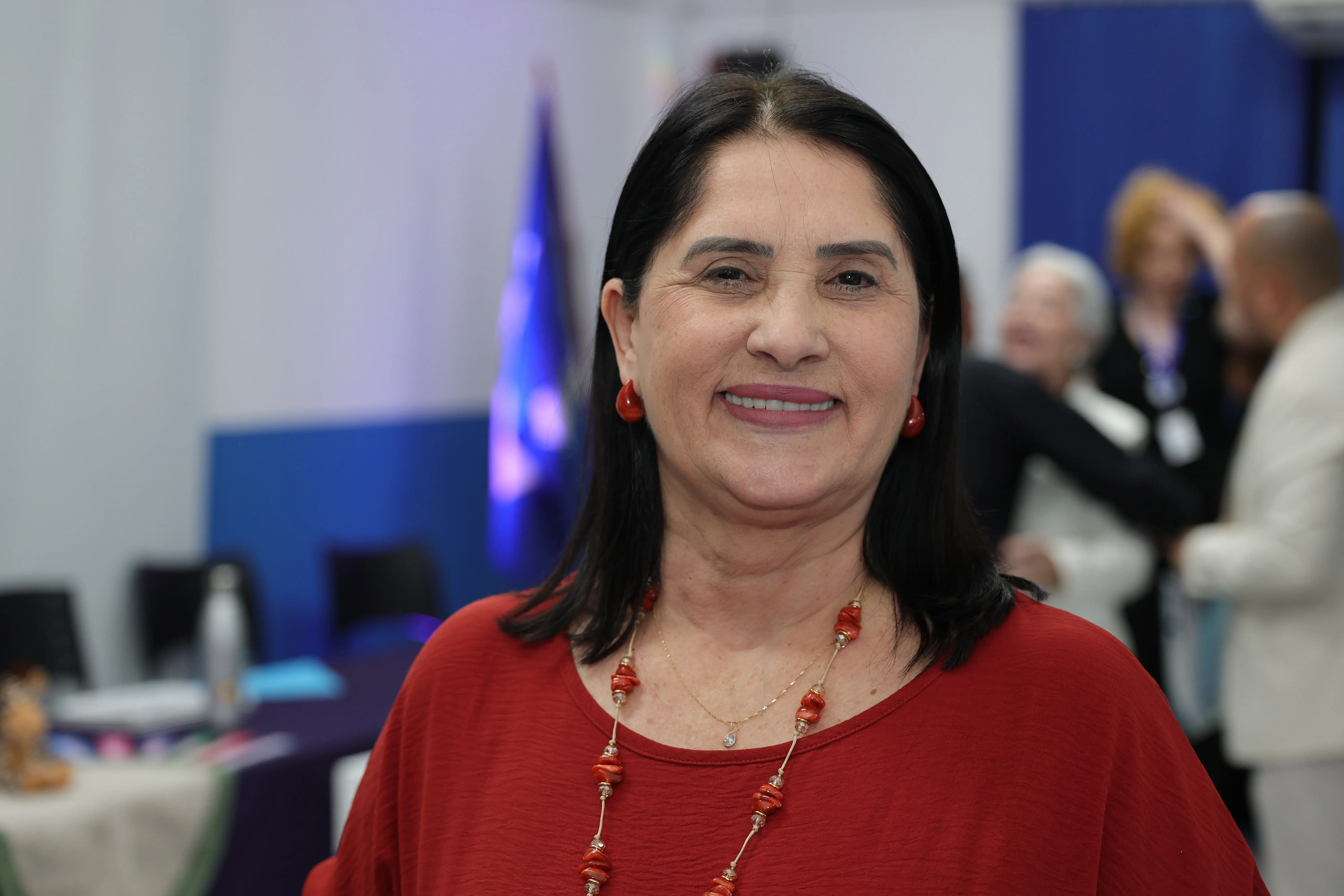 Mulher de cabelos escuros, blusa vermelha e colar de contas na mesma cor, sorri em ambiente de evento. Ao fundo, pessoas confraternizam próximas a um painel azul iluminado.