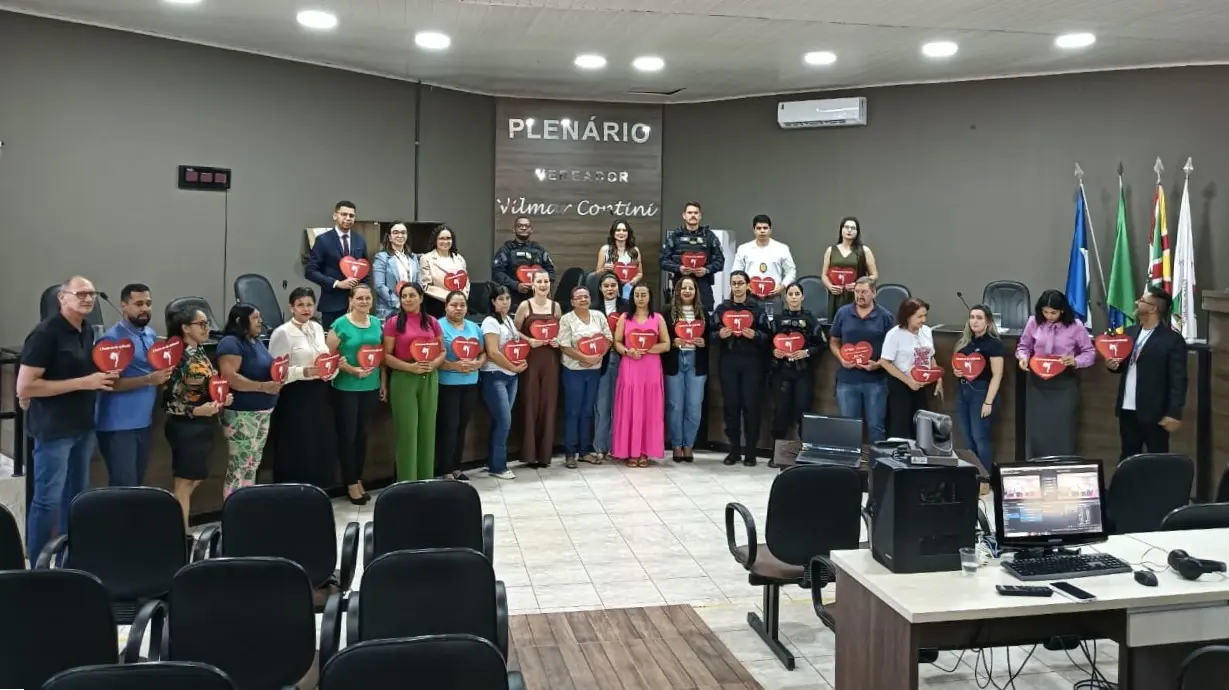 Grupo de pessoas em pé no plenário de uma câmara municipal segura placas vermelhas em formato de coração. Ao fundo, pare de madeira com a inscrição 