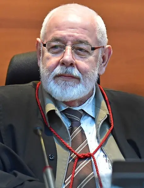Foto vertical que mostra o presidente do TJMT, desembargador José Zuquim Nogueira, sentado em seu posto no plenário do tribunal, olhando para o lado. Ele é um senhor branco, de cabelo e barba brancos, usando toga e óculos de grau.